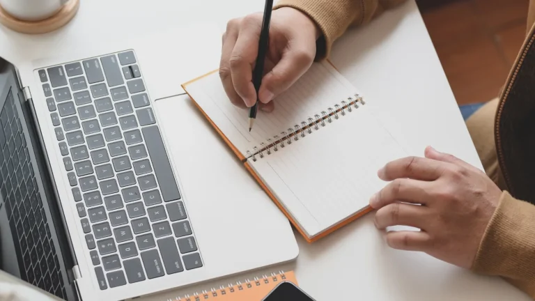 Person taking notes by hand while working on a laptop, representing training and evidence review